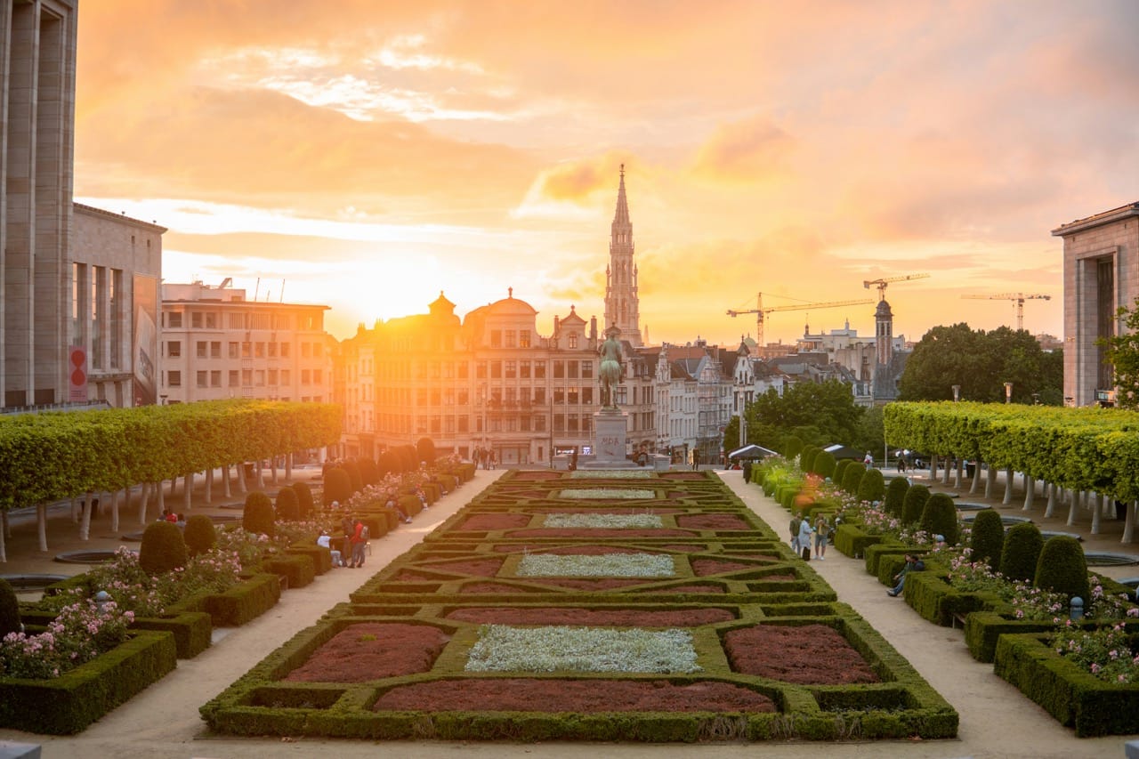 Vue panoramique du Mont des Arts à Bruxelles, Belgique, avec ses jardins bien entretenus, des bâtiments historiques en arrière-plan et un coucher de soleil spectaculaire illuminant la scène.