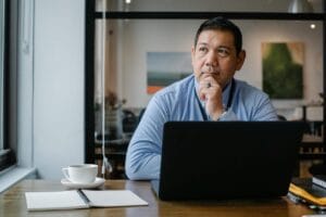 Homme en chemise bleue assis à son bureau, réfléchissant devant son ordinateur portable, avec une tasse de café et un carnet ouverts, dans un bureau moderne avec une ambiance professionnelle.