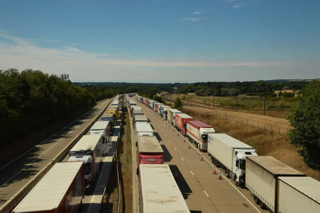 Vue aérienne d’une file ininterrompue de camions à l’arrêt sur une autoroute, déviés sur une seule voie, dans un paysage rural sous ciel dégagé.